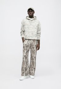 Felpa con cappuccio e pantaloni a gamba larga in beige tenue con un motivo a foglie. Il tessuto sembra morbido, con una vestibilità comoda. Sneakers bianche completano il look.