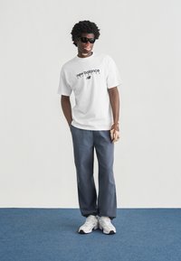 Άσπρο βαμβακερό t-shirt με το λογότυπο "new balance", γκρι φαρδιά παντελόνια και άσπρα αθλητικά παπούτσια με γκρι λεπτομέρειες, όρθια σε μπλε χαλί.