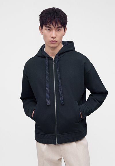 Νέος άνδρας φορώντας μαύρο hoodie με φερμουάρ και κορδόνια και μπεζ παντελόνι, στέκεται με τα χέρια στις τσέπες σε απλό φόντο.