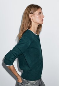 Türkisfarbener Wollcardigan mit rundem Ausschnitt, Knöpfen vorne und gerippten Bündchen. Stylisch kombiniert mit hellfarbenen langen Ärmeln und grauen Jeans.