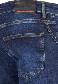 Gros plan de la poche arrière d'un jean en denim bleu avec une couture orange et une chaîne métallique attachée à une boucle de ceinture.