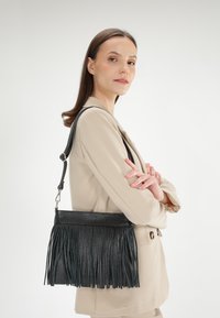 Černá kožená kabelka s třásněmi, obdélníkového tvaru a nastavitelným popruhem, nošená přes světle béžový outfit. Hladká textura, minimalistický design.