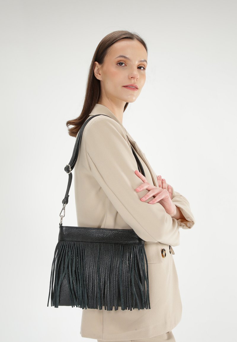 Černá kožená kabelka s třásněmi, obdélníkového tvaru a nastavitelným popruhem, nošená přes světle béžový outfit. Hladká textura, minimalistický design.