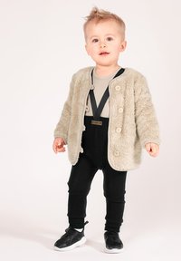 Miękki beżowy kardigan z dużymi guzikami, nałożony na szary prążkowany top, czarne szelki oraz prążkowane czarne spodnie; dopełnieniem stroju są czarne trampki.