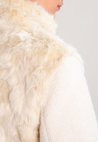 Faux-Fur-Weste in Creme mit weicher, flauschiger Textur. Auf einer Seite aus weißem Pullovermaterial, nahtloser Übergang zwischen den Stoffen.