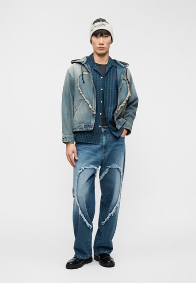 Veste en jean bleu avec bords effilochés, chemise à carreaux, jean déchiré avec des découpes en forme de cœur, et un bonnet de couleur claire.