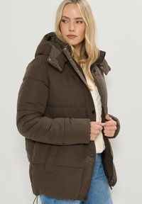 Brun vadderad jacka med avtagbar huva, tryckknapp- och dragkedjestängning, samt två sidofickor. Texturerat tyg med synligt kviltat mönster.