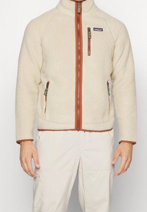 Beige fleecetakjakke med rustrøde lynlås- og lommekanter, med en brystlomme og to sidelommer, båret med lysebeige bukser.