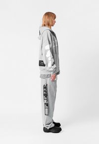 Sudadera con capucha gris de algodón con gran texto blanco, combinada con pantalones de chándal grises a juego que presentan detalles gráficos en negro y bolsillos de carga.