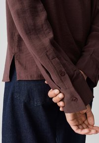 Person wearing dark brown long-sleeve shirt with buttoned cuffs holding wrist behind their back, paired with dark blue denim pants.