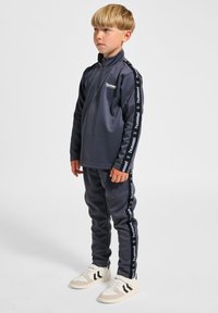 Marineblauer Zip-up-Trainingsanzug mit schwarzen, logoed Seitenschreifen, hoher Kragen und figurbetontem Design; kombiniert mit weißen Sneakern.