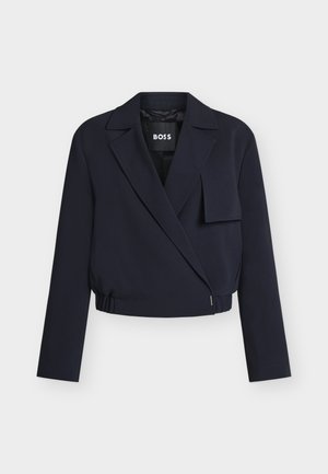 Blazer cropped azul marino con solapas anchas, cierre frontal asimétrico, mangas largas y una solapa decorativa de tela en el pecho derecho.