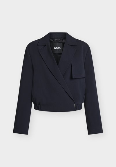 Blazer court bleu marine avec larges revers, fermeture asymétrique à l'avant, manches longues et rabat décoratif en tissu sur la poitrine droite.