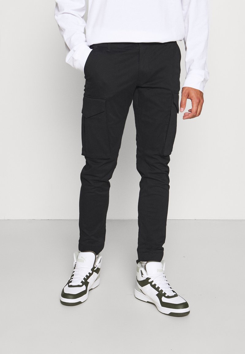 Zwarte cargobroek met een slim fit, voorzien van voor- en zijzakken, gemaakt van een duurzaam materiaal. Gecombineerd met wit en groen hoge sneakers.