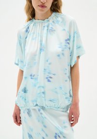 Ljust blå blus i mjuk tyg, med en sammetslen halsringning, korta ärmar och subtila abstrakta mönster i mörkare blå nyanser.