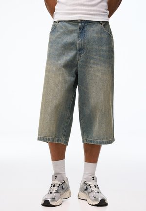 Férfi, elhasznált, bő farmer rövidnadrágban, fehér zokniban, ezüst sportcipővel, sima fehér háttér előtt.