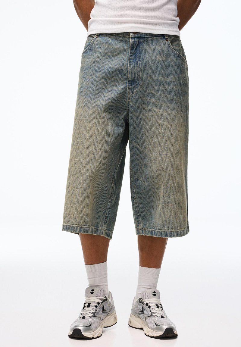 Homme portant un short en jean délavé et ample, des chaussettes blanches et des baskets argentées sur un fond blanc uni.