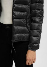 Zwarte pufferjack met een gequiltde textuur, ritssluiting en elastische manchetten. Gecombineerd met een crèmekleurige top en zwarte jeans, wat een gelaagde look toont.
