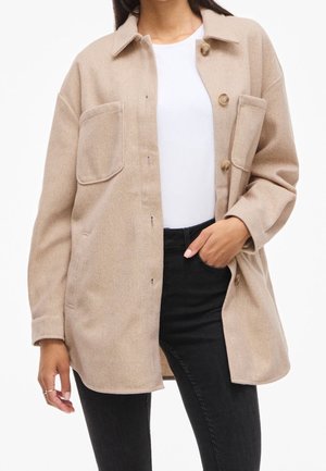 Kvinde iført en beige jakke med knapper og store lommer, en hvid skjorte og sorte bukser, med den ene hånd i lommen.