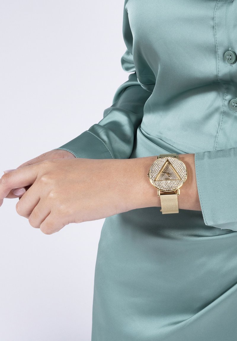 Reloj de pulsera dorado con cara triangular, adornado con cristales y una correa de malla metálica; combinado con una camisa de satén verde de manga larga.