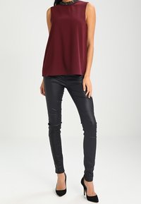 Haut sans manches bourgogne avec un col en perles, associé à un pantalon en cuir synthétique noir et des escarpins noirs. Texture lisse, coupe ajustée.