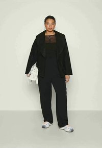 Black blazer with a large collar over a sheer black top, paired with wide-leg black pants and light sneakers. Accessorized with a white handbag.