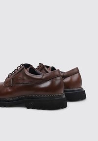Zapatos de cuero marrón con una textura suave, forma redondeada y suelas negras resistentes en contraste. Presentan ojales y acentos perforados.