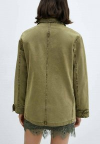 Osoba s hnědými vlasy po ramenou má na sobě olivově zelenou bundu přes tmavě zelenou krajkovou sukni, viděná zepředu na neutrálním pozadí.