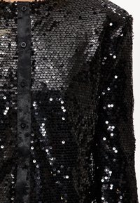 Veste noire en sequins avec une fermeture avant à boutons. Le tissu est orné de sequins brillants superposés, créant une surface texturée et réfléchissante.