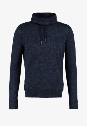 Mies pukeutuneena tummansiniseen neulepaitaan, jossa on nyörit kauluksessa, ja haalistuneisiin sinisiin farkkuihin, käsi taskussa, yksinkertaista vaaleaa taustaa vasten.