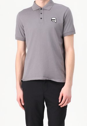 Mann trägt ein graues Kurzarm-Poloshirt mit einem kleinen schwarz-weißen Emoji-Aufnäher auf der linken Brust, kombiniert mit schwarzen Hosen.