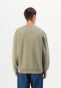 Sudadera de cuello redondo en verde oliva con hombros caídos, puños y dobladillo de canalé, y una textura suave. Combinada con unos vaqueros azules de corte holgado.