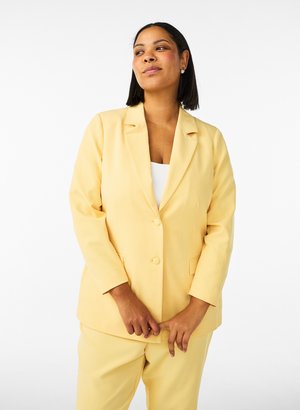 Donna con capelli lisci e scuri che indossa orecchini di perle e un completo giallo, in piedi davanti a uno sfondo bianco uniforme.