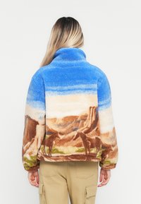Personne portant une veste sherpa avec un paysage désertique et un design de cheval à l'arrière, associée à un pantalon cargo beige.
