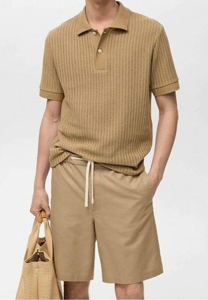 Homme portant un polo en maille beige et un short assorti, tenant un sac fourre-tout tissé beige dans une main, l’autre main dans la poche.