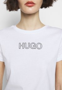 Camiseta de algodón blanca con mangas cortas y cuello redondo. "HUGO" impreso en negrita en color negro en el pecho. Textura suave.