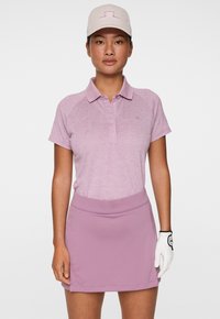 Donna che indossa una polo da golf di un leggero viola, una gonna abbinata, un cappello chiaro e un guanto da golf bianco sulla mano destra, in piedi contro uno sfondo bianco.