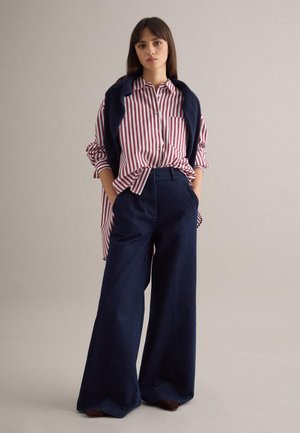 Frau steht mit den Händen in den Taschen, trägt ein rot-weiß gestreiftes Hemd, marineblaue Hosen mit weitem Bein und einen marineblauen Pullover, der über den Schultern liegt.