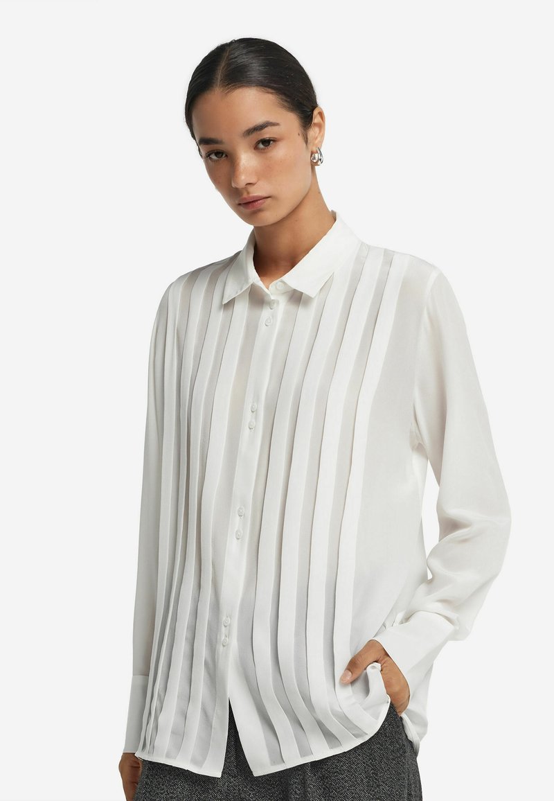 Camicia bianca con bottoni, caratterizzata da pieghe verticali, tessuto morbido, maniche lunghe e colletto classico. Leggera lucentezza e vestibilità rilassata.