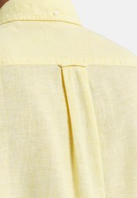 Lichtgele linnen shirt met een rugplooitje en kleine knoopdetails bij de kraag. Zachte textuur met een licht doorzichtige uitstraling.