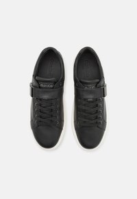 Baskets en cuir noir avec un design à lacets, une sangle ajustable et une semelle en caoutchouc blanc. La marque est discrètement embossée sur la semelle intérieure.