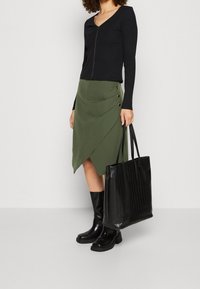 Top nero a coste con zip dorata, gonna avvolgente verde con bottoni laterali, stivali neri alti e una borsa a pieghe nera.