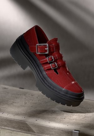 Chaussure en cuir rouge brillant avec trois boucles argentées et semelle épaisse noire sur une surface en béton avec un fond gris.