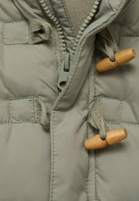 Beige vuorattu takki, jossa on suljettu muovivetoketju ja kaksi puutappi kiinnitystä harmailla kankaisilla lenkeillä.