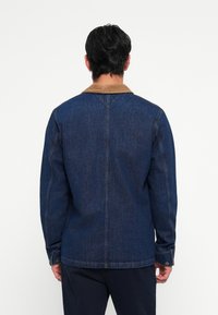 Chaqueta de denim azul oscuro con un cuello de pana marrón y mangas largas; presenta costuras verticales y puños abotonados.
