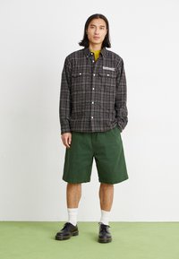 Chemise à carreaux noir et blanc à boutons avec deux poches poitrine, associée à un short vert et des chaussures noires, portée avec des chaussettes blanches.