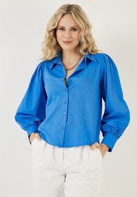 Bright blue button-up shirt with a collared neckline and puff sleeves. Smooth fabric with a relaxed fit and cuffed wrists.