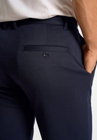 Navy trousers made of smooth fabric featuring a single rear pocket with a black button detail. Tailored design with a slim fit.
