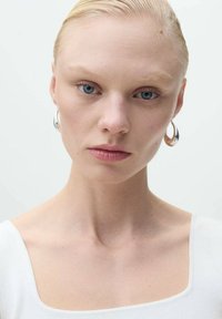 Modelka s bledou pletí a blond vlasy, oblečená v bílém topu. Mezi její výrazné rysy patří modré oči a stříbrné kruhové náušnice.