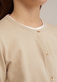 Bežs kardigans ar apaļu kaklu, ar zelta pogām un ribota tekstūra. Uzvilkts pār baltu krekla, parādot slāņveida stilu.